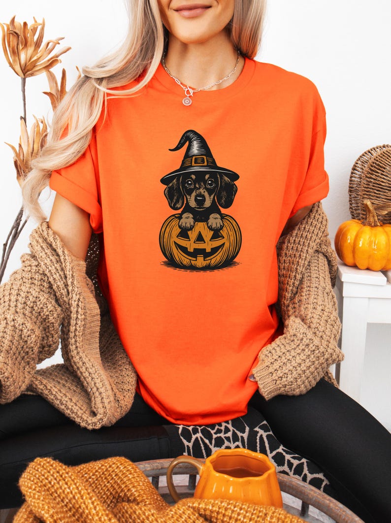 May include: Orange t-shirt featuring a black and tan dachshund wearing a witch hat, sitting inside a carved jack-o'-lantern. The design is a Halloween theme. The shirt is made of a soft material.