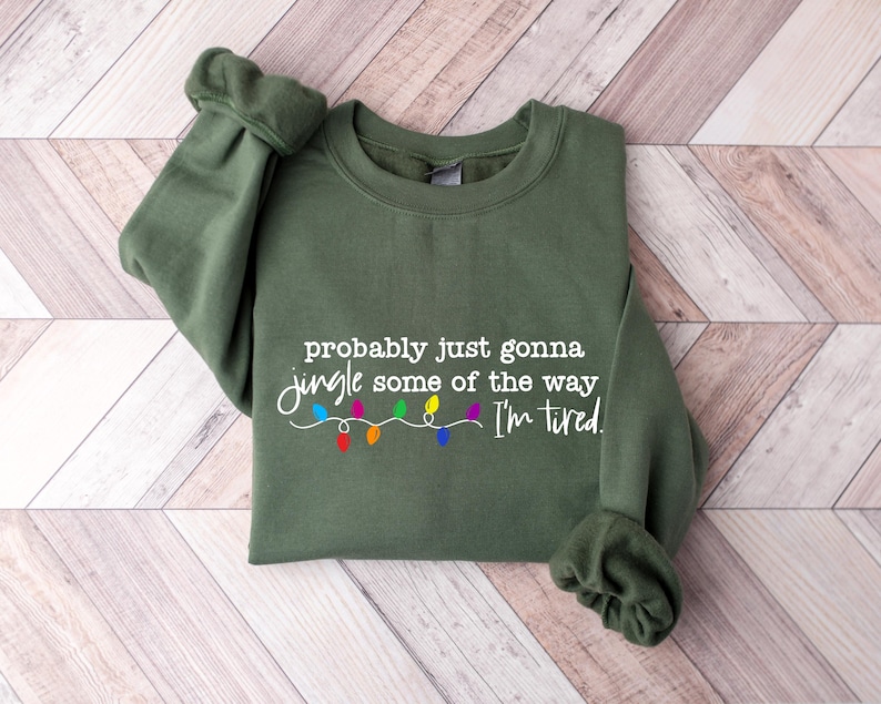Puede incluir: Una sudadera verde oliva con el texto "probably just gonna jingle some of the way I'm tired" en escritura blanca. Debajo, una cadena de luces navide&ntilde;as de colores. La sudadera est&aacute; doblada sobre una superficie de madera clara.