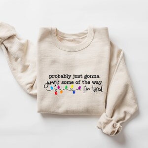 Puede incluir: Sudadera beige con la frase "probably just gonna jingle some of the way I'm tired" en escritura negra. Debajo, una cadena de luces navide&ntilde;as de colores. Las mangas est&aacute;n dobladas.