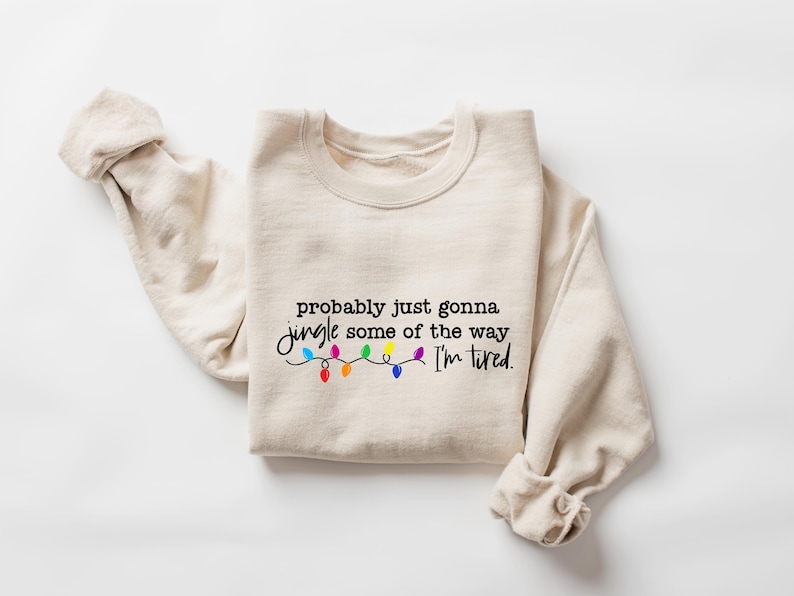 Puede incluir: Sudadera beige con la frase "probably just gonna jingle some of the way I'm tired" en escritura negra. Debajo, una cadena de luces navide&ntilde;as de colores. Las mangas est&aacute;n dobladas.
