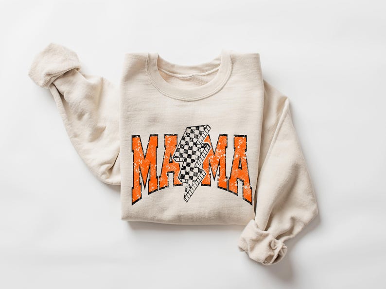 Pu&ograve; includere: Felpa color crema con la scritta "MAMA" in lettere arancioni sbiadite. Un fulmine a scacchi in bianco e nero &egrave; centrato sopra la parola. Le maniche sono piegate.