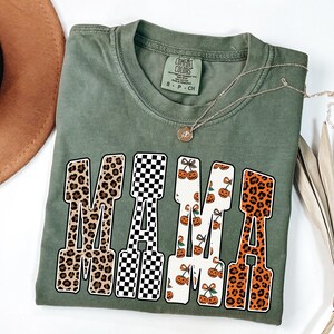 May include: An olive green t-shirt with the word "MAMA" in a bold, block letter design. The letters feature various patterns, including leopard print, a black and white checkerboard, and a pumpkin and cherry design. A gold necklace rests on the shirt.