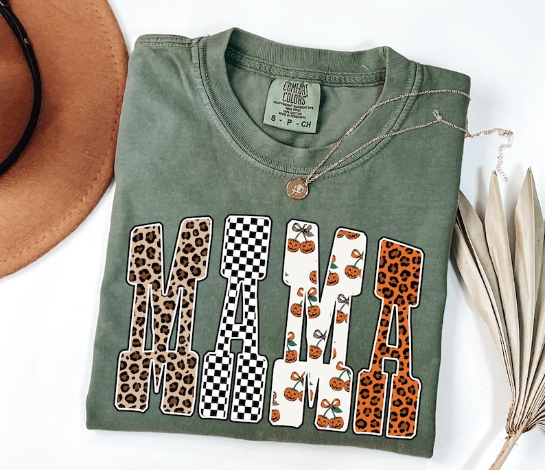 May include: An olive green t-shirt with the word "MAMA" in a bold, block letter design. The letters feature various patterns, including leopard print, a black and white checkerboard, and a pumpkin and cherry design. A gold necklace rests on the shirt.