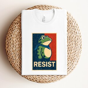 May include: White t-shirt featuring a cartoon frog with a blue scarf, set against a red and blue background. The word "RESIST" is printed in bold letters below the frog. The shirt is folded on a woven surface.