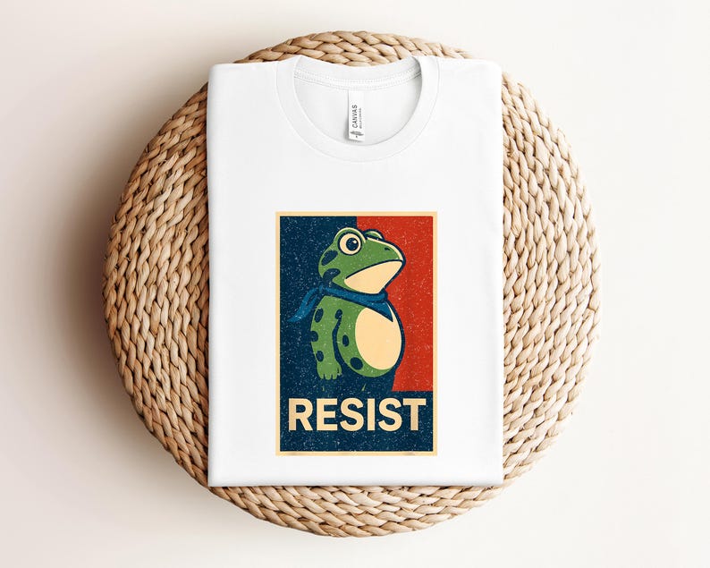 May include: White t-shirt featuring a cartoon frog with a blue scarf, set against a red and blue background. The word "RESIST" is printed in bold letters below the frog. The shirt is folded on a woven surface.