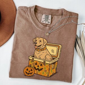 Puede incluir: Una camiseta Comfort Colors marr&oacute;n con un dibujo animado de un perro Labrador Retriever en una nevera y una calabaza de Halloween. El perro lleva un collar. Un collar plateado est&aacute; en la camiseta. La camiseta es talla S.