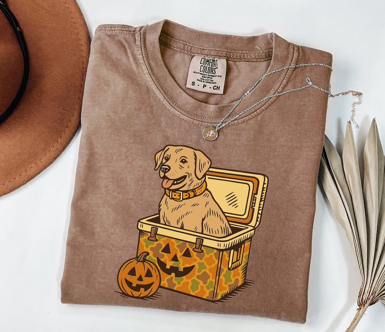 Puede incluir: Una camiseta Comfort Colors marr&oacute;n con un dibujo animado de un perro Labrador Retriever en una nevera y una calabaza de Halloween. El perro lleva un collar. Un collar plateado est&aacute; en la camiseta. La camiseta es talla S.