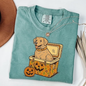 Puede incluir: Una camiseta Comfort Colors verde azulado con un perro Labrador Retriever de dibujos animados sentado en una nevera y una calabaza. La nevera tiene un patr&oacute;n de camuflaje. Un collar plateado est&aacute; sobre la camisa.