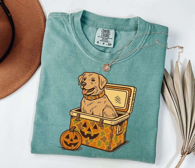 Puede incluir: Una camiseta Comfort Colors verde azulado con un perro Labrador Retriever de dibujos animados sentado en una nevera y una calabaza. La nevera tiene un patr&oacute;n de camuflaje. Un collar plateado est&aacute; sobre la camisa.