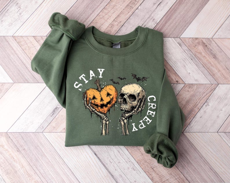May include: A green sweatshirt with the words "STAY CREEPY" in white. The graphic features a jack-o'-lantern heart held by skeletal hands next to a skull, with bats above. The sweatshirt is folded on a wooden surface.