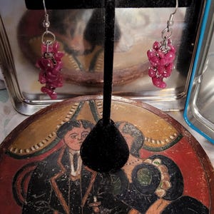 May include: A pair of dangle earrings with a cluster of small, irregular, dark pink beads. The beads are strung together with silver-colored wire and hang from a silver-colored hook. The earrings are displayed on a black velvet earring stand.