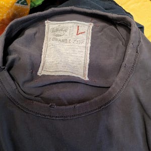 Może przedstawiać: Zbliżenie na ciemnoszarą koszulkę z postarzanym dekoltem. Prostokątna metka z tekstem i grafiką korony jest wszyta z tyłu koszulki. Na metce znajduje się napis "Z-BRAND Z-ブランド".
