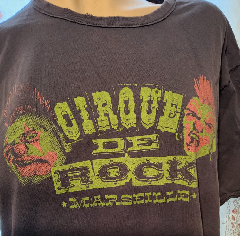 Może przedstawiać: Ciemnobrązowa koszulka z napisem "CIRQUE DE ROCK MARSEILLE" w kolorze zielonym i ż&oacute;łtym. Koszulka posiada dwie grafiki postaci punk rockowych z zielonymi i czerwonymi akcentami. Koszulka ma postarzany wygląd.