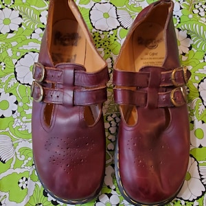May include: A pair of burgundy leather Mary Jane shoes with two buckle straps and decorative perforations. The shoes have a round toe and a black sole with yellow stitching. The brand name is visible inside the shoe.