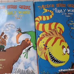 May include: Two hardcover books, "The Early Works of Dr. Seuss," Volume 1 and 2, are displayed. Volume 1 features a yellow fish with red stripes, while Volume 2 shows cartoon figures and text. The covers are blue and have the title "Theodor Seuss Geisel."
