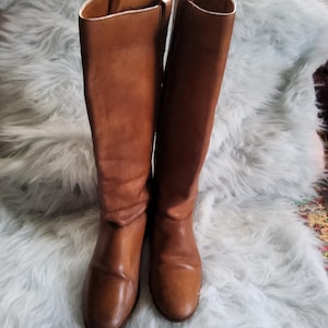 May include: A pair of knee-high brown leather boots. The boots have a classic design with a rounded toe and a slight heel. The leather has a smooth texture and a warm, medium brown color. The boots are set against a light blue, fluffy background.