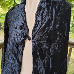 May include: A black crushed velvet jacket featuring long sleeves and a single button closure at the center front. The garment has a textured, crinkled surface finish and is displayed on a mannequin, showing a draped, open-front silhouette.