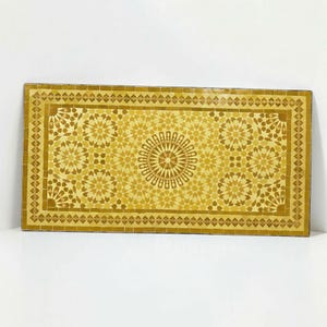 K&ouml;nnte beinhalten: Eine rechteckige Tischplatte mit einer Mosaikoberfl&auml;che aus kleinen, handgeschnittenen Fliesen in verschiedenen Gelb- und Goldt&ouml;nen. Das Design zeigt ein zentrales Sternmuster, umgeben von sich wiederholenden geometrischen Motiven und einer Umrandung.