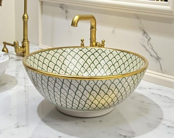 May include: A decorative, round bathroom sink with a white and green patterned design. The sink has a gold rim and is paired with a gold faucet. The sink is a vessel sink, sitting on a marble countertop.