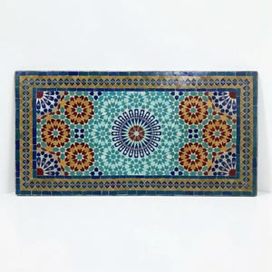 K&ouml;nnte beinhalten: Eine rechteckige Tischplatte mit einem komplexen Mosaikmuster aus handgeschnittenen Fliesen in T&uuml;rkis, Dunkelblau, Senfgelb und Dunkelrot. Das Design zeigt ein zentrales Sternmotiv, das von sich wiederholenden geometrischen Blumenformen umgeben ist.