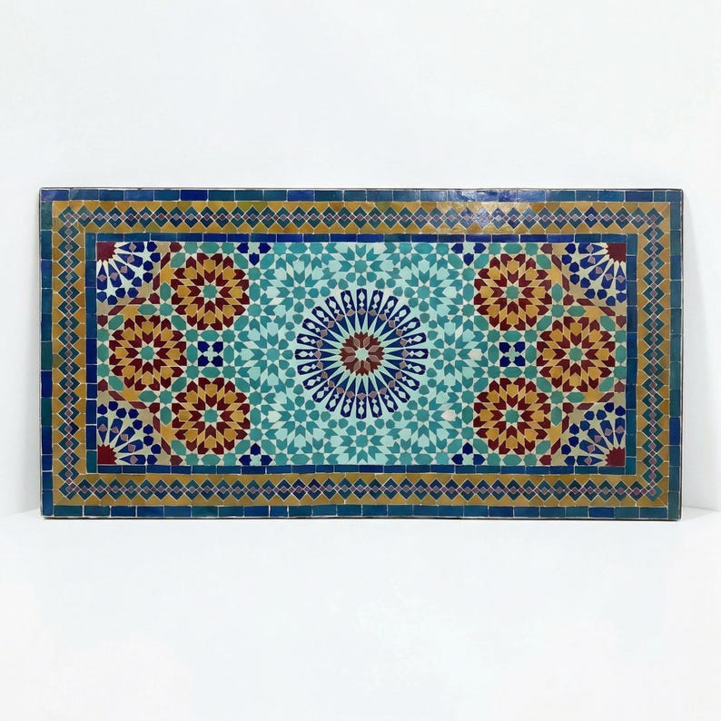 K&ouml;nnte beinhalten: Eine rechteckige Tischplatte mit einem komplexen Mosaikmuster aus handgeschnittenen Fliesen in T&uuml;rkis, Dunkelblau, Senfgelb und Dunkelrot. Das Design zeigt ein zentrales Sternmotiv, das von sich wiederholenden geometrischen Blumenformen umgeben ist.