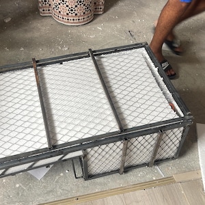 Puede incluir: Un marco met&aacute;lico rectangular con una parte superior de malla de alambre y material blanco en el interior. El marco est&aacute; sobre un suelo de hormig&oacute;n. Una persona est&aacute; de pie cerca.