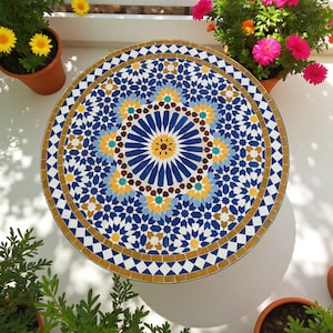 Może przedstawiać: Okrągły stół z mozaiki z bogatym geometrycznym wzorem w odcieniach niebieskiego, żółtego i brązowego. Krawędź stołu ma złoty obramowanie. Stół otaczają rośliny doniczkowe z kolorowymi kwiatami.