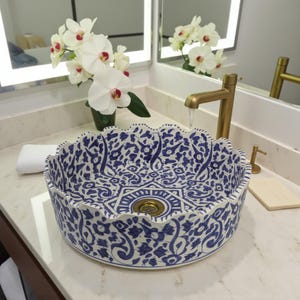 Puede incluir: Un lavabo redondo de cerámica decorativo con un patrón floral azul y blanco. El lavabo tiene un borde festoneado y un desagüe de latón. Se ve un grifo de latón, con agua fluyendo hacia el lavabo. Orquídeas blancas en el fondo.
