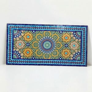 K&ouml;nnte beinhalten: Eine rechteckige Mosaikfliesenplatte mit einem komplexen geometrischen Muster. Das Design umfasst ein zentrales kreisf&ouml;rmiges Medaillon, das von sich wiederholenden sternf&ouml;rmigen Motiven in den Farben Dunkelblau, T&uuml;rkis, Olivgr&uuml;n und Goldgelb umgeben ist.