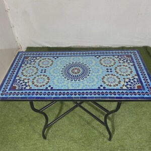 K&ouml;nnte beinhalten: Ein rechteckiger Tisch mit einer Mosaik-Tischplatte, die ein geometrisches Muster in Blau-, Hellblau- und Wei&szlig;t&ouml;nen aufweist. Der Tisch wird von einem schwarzen Metallrahmen mit vier geschwungenen Beinen getragen, die eine X-f&ouml;rmige Basis bilden.