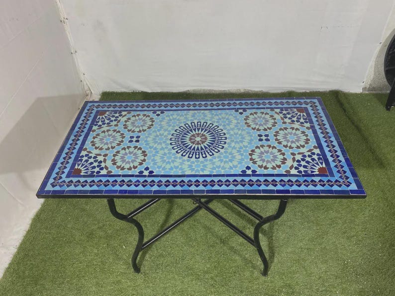 K&ouml;nnte beinhalten: Ein rechteckiger Tisch mit einer Mosaik-Tischplatte, die ein geometrisches Muster in Blau-, Hellblau- und Wei&szlig;t&ouml;nen aufweist. Der Tisch wird von einem schwarzen Metallrahmen mit vier geschwungenen Beinen getragen, die eine X-f&ouml;rmige Basis bilden.