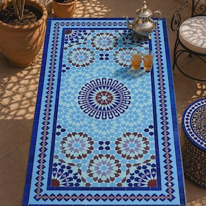 K&ouml;nnte beinhalten: Ein rechteckiger Tisch mit einer Mosaik-Tischplatte mit komplexen geometrischen Mustern in Hellblau, Dunkelblau und Braun. Der Tisch steht auf einem schwarzen Metallgestell mit geschwungenen Beinen und ist mit einer silbernen Teekanne und zwei Gl&auml;sern versehen.