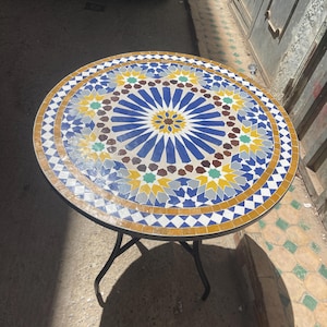 May include: Circular mosaic tile table with a black metal frame. The tabletop has a detailed geometric pattern in blue, white, yellow, and brown tiles. The table is approximately 76 cm in diameter.
