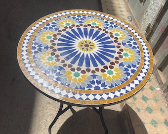 Könnte beinhalten: Runder Mosaiktisch mit schwarzem Metallrahmen. Die Tischplatte hat ein detailliertes geometrisches Muster aus blauen, weißen, gelben und braunen Fliesen. Der Tisch hat einen Durchmesser von ca. 76 cm.