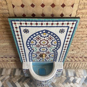 Puede incluir: Una fuente decorativa de mosaico con una pila turquesa y un intrincado trabajo de azulejos. La fuente presenta una boquilla de lat&oacute;n y est&aacute; situada sobre un fondo de ladrillos y azulejos estampados. El dise&ntilde;o del mosaico incluye patrones geom&eacute;tricos azules, blancos, rojos y amarillos.
