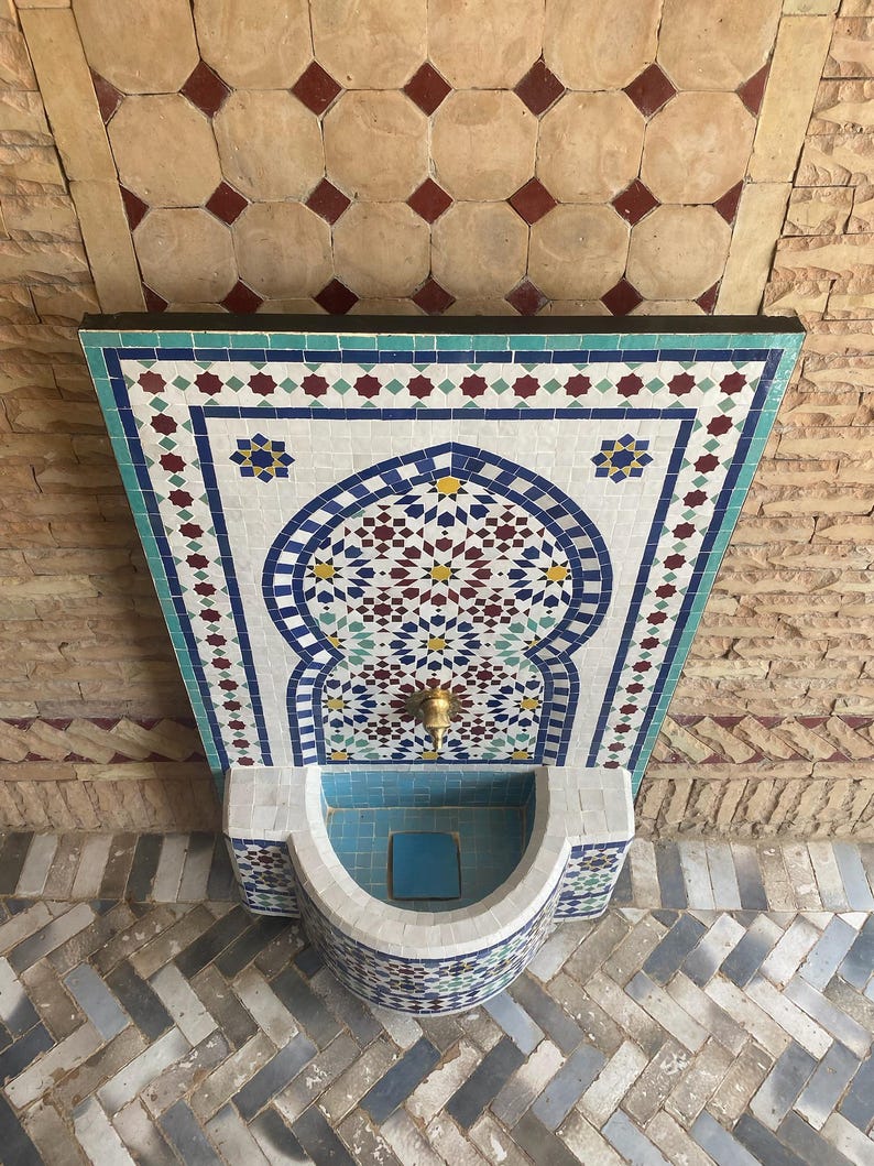 Puede incluir: Una fuente decorativa de mosaico con una pila turquesa y un intrincado trabajo de azulejos. La fuente presenta una boquilla de lat&oacute;n y est&aacute; situada sobre un fondo de ladrillos y azulejos estampados. El dise&ntilde;o del mosaico incluye patrones geom&eacute;tricos azules, blancos, rojos y amarillos.