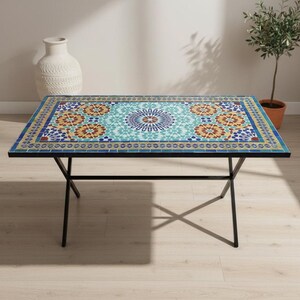 K&ouml;nnte beinhalten: Ein rechteckiger Tisch mit einer Mosaik-Tischplatte, die ein zentrales kreisf&ouml;rmiges Medaillon und sich wiederholende florale Muster in T&uuml;rkis, Gelb, Orange und Blau aufweist, getragen von einem schwarzen Metallrahmen mit d&uuml;nnen, gekreuzten Beinen.