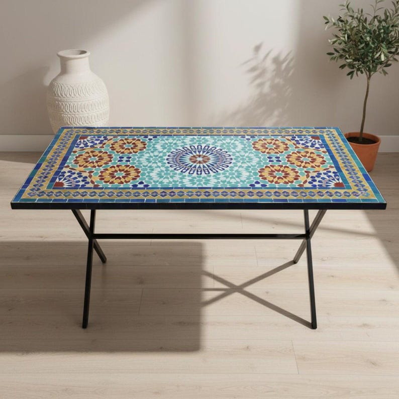 K&ouml;nnte beinhalten: Ein rechteckiger Tisch mit einer Mosaik-Tischplatte, die ein zentrales kreisf&ouml;rmiges Medaillon und sich wiederholende florale Muster in T&uuml;rkis, Gelb, Orange und Blau aufweist, getragen von einem schwarzen Metallrahmen mit d&uuml;nnen, gekreuzten Beinen.