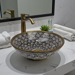 Peut inclure: Un lavabo décoratif avec un intérieur à motifs noirs et blancs et un bord doré. Le lavabo est associé à un robinet doré et repose sur un comptoir blanc. Un miroir et des serviettes sont en arrière-plan.