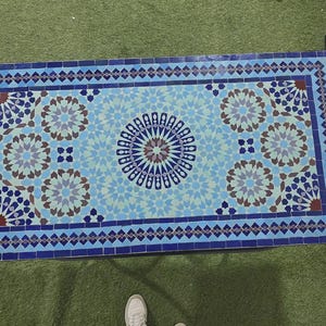 K&ouml;nnte beinhalten: Eine rechteckige Tischplatte mit einem Mosaikfliesenmuster in Blau-, Hellblau- und Braunt&ouml;nen. Das Design besteht aus einem zentralen Sternmotiv, das von sich wiederholenden geometrischen Blumenmustern umgeben und von einem dunkelblauen Rand eingefasst ist.
