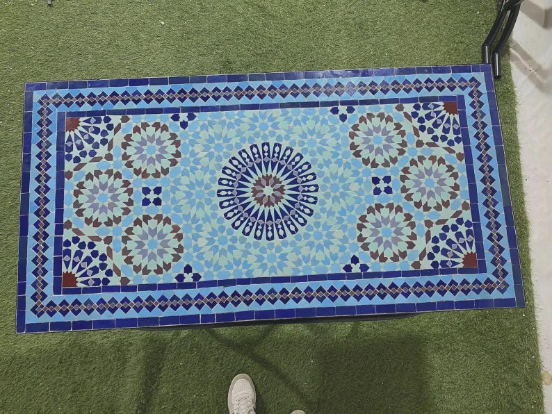 K&ouml;nnte beinhalten: Eine rechteckige Tischplatte mit einem Mosaikfliesenmuster in Blau-, Hellblau- und Braunt&ouml;nen. Das Design besteht aus einem zentralen Sternmotiv, das von sich wiederholenden geometrischen Blumenmustern umgeben und von einem dunkelblauen Rand eingefasst ist.