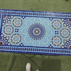 K&ouml;nnte beinhalten: Eine rechteckige Tischplatte mit einem Mosaikfliesenmuster in Blau-, Hellblau- und Braunt&ouml;nen. Das Design umfasst ein zentrales Sternmotiv, das von sich wiederholenden geometrischen Blumenmustern und einem dunkelblauen Rahmen umgeben ist.