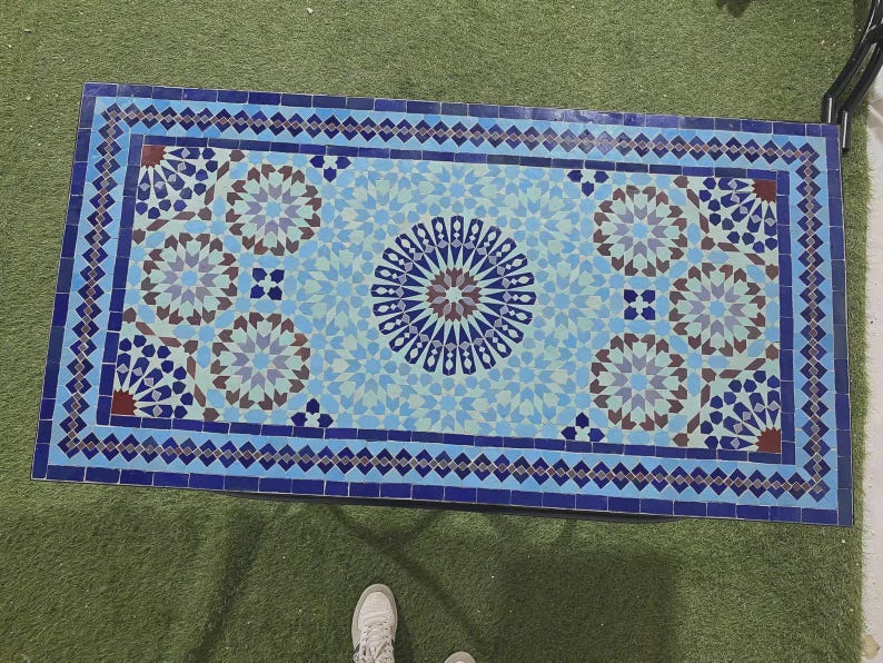 K&ouml;nnte beinhalten: Eine rechteckige Tischplatte mit einem Mosaikfliesenmuster in Blau-, Hellblau- und Braunt&ouml;nen. Das Design umfasst ein zentrales Sternmotiv, das von sich wiederholenden geometrischen Blumenmustern und einem dunkelblauen Rahmen umgeben ist.