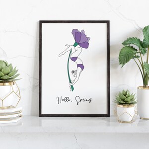 Könnte beinhalten: Ein gerahmter Kunstdruck mit einer Strichzeichnung einer Person, die an einer Blume hängt. Die Person trägt lila Badebekleidung. Der Text "Hello, Spring" steht unter dem Bild. Der Druck wird auf einer weißen Oberfläche mit Pflanzen ausgestellt.