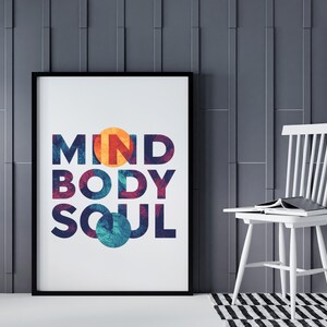 May include: A framed print displaying the words "MIND BODY SOUL" in vibrant colours. The artwork is set against a grey wall, with a white chair and a black and white rug in the background. The frame is black.