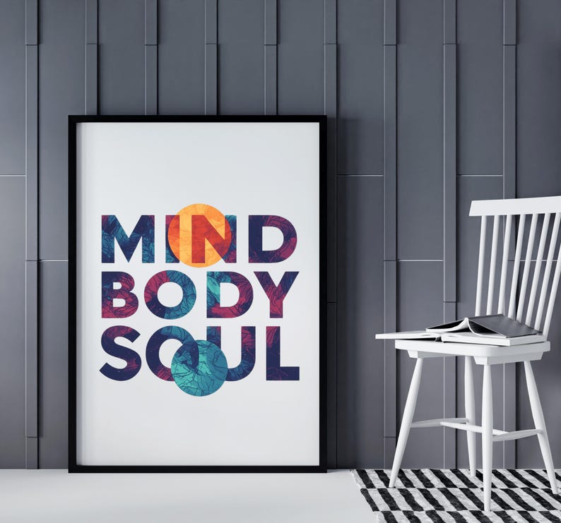 May include: A framed print displaying the words "MIND BODY SOUL" in vibrant colours. The artwork is set against a grey wall, with a white chair and a black and white rug in the background. The frame is black.