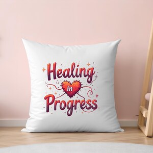 May include: White square cushion featuring the phrase "Healing in Progress" in a decorative font. The design includes a red heart with a stitched border and a small "in" inside the heart. The background is a light pink.