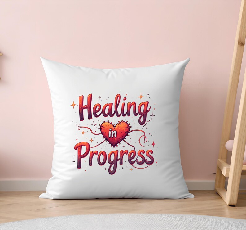 May include: White square cushion featuring the phrase "Healing in Progress" in a decorative font. The design includes a red heart with a stitched border and a small "in" inside the heart. The background is a light pink.