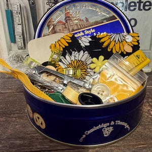 Puede incluir: Una lata azul de kit de costura Cambridge & Thames llena de suministros de costura. La lata contiene tela, botones, hilo y una cinta métrica amarilla. La tapa presenta una ilustración de un castillo. Otras herramientas de costura son visibles en el fondo.