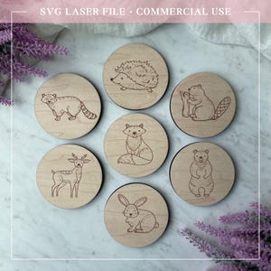 Peut inclure: Un ensemble de sept dessous de verre en bois avec des motifs d'animaux gravés au laser, dont un raton laveur, un hérisson, un castor, un renard, un cerf, un ours et un lapin. Les dessous de verre sont ronds et en bois clair. Le texte "SVG LASER FILE + COMMERCIAL USE" est en haut.